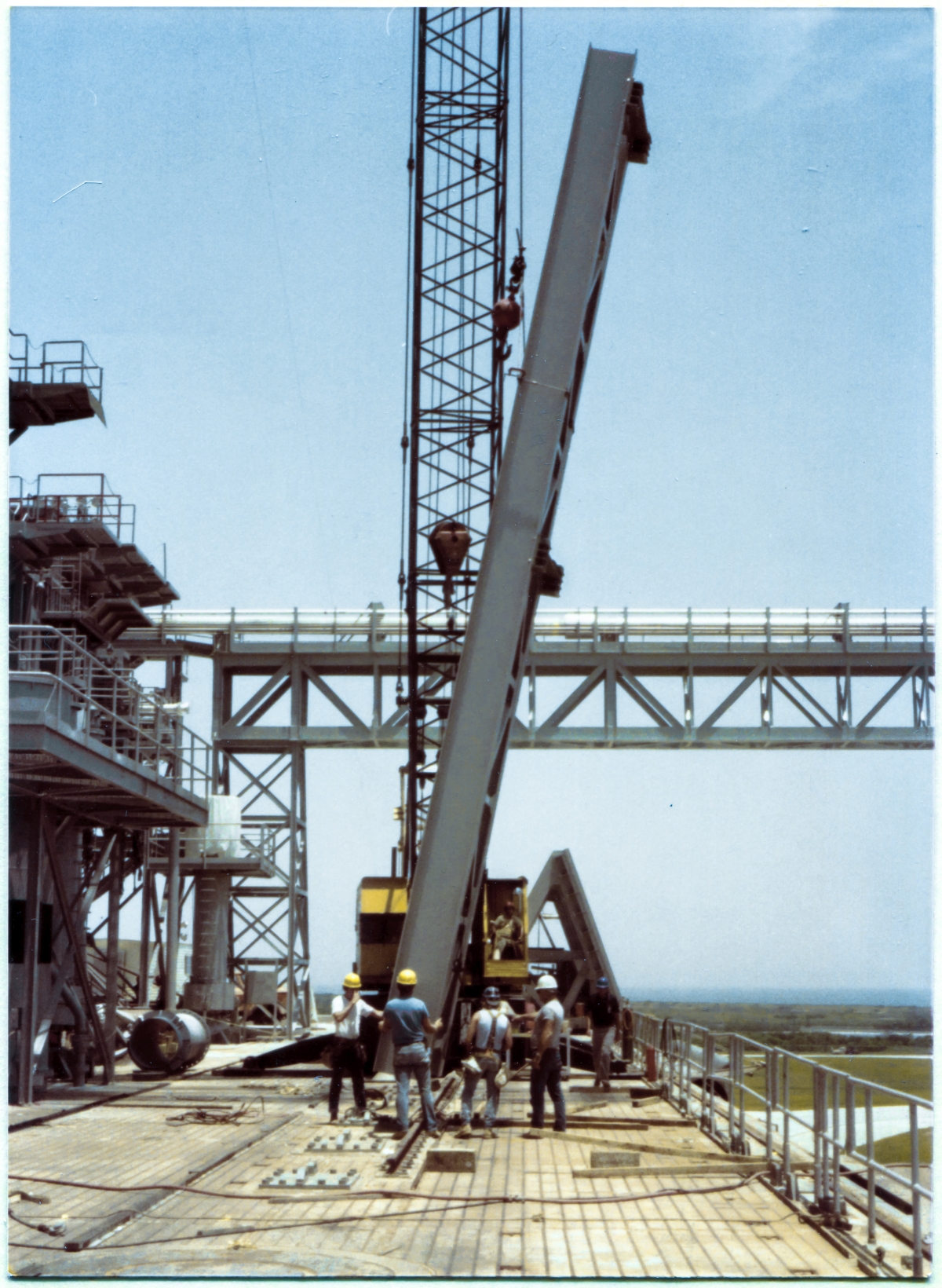 Image 077. The GOX Arm Strongback remains suspended just above the Pad Deck, still within easy reach, as our small group works out the precise arrangements which will need to be effected from here on, as the Strongback rises above the ground, far out of reach, until it arrives at its destination on the Fixed Service Structure, 250 feet straight up from the Pad Deck it now hangs less than a yard above. For the moment, it is rotated 180 degrees away from its final orientation, flush against the Primary Framing on side 1 of the FSS, extending from elevation 260'-0” to 300'-0”. During lifts, orientations of that which is being lifted do not necessarily match their final orientations, and this can be for any number of reasons, most of which are only apparent to those persons directly involved in the lift, and none of which might be visually apparent to an untrained or uninvolved observer. Photo by James MacLaren.